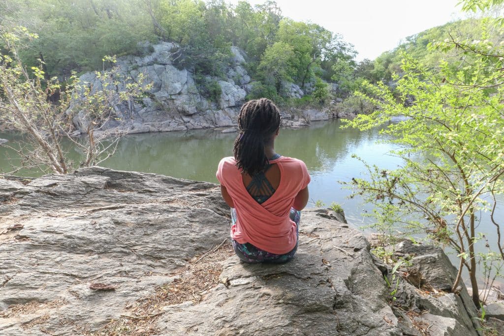 Mulher negra sentada em uma rocha em frente a um lago.