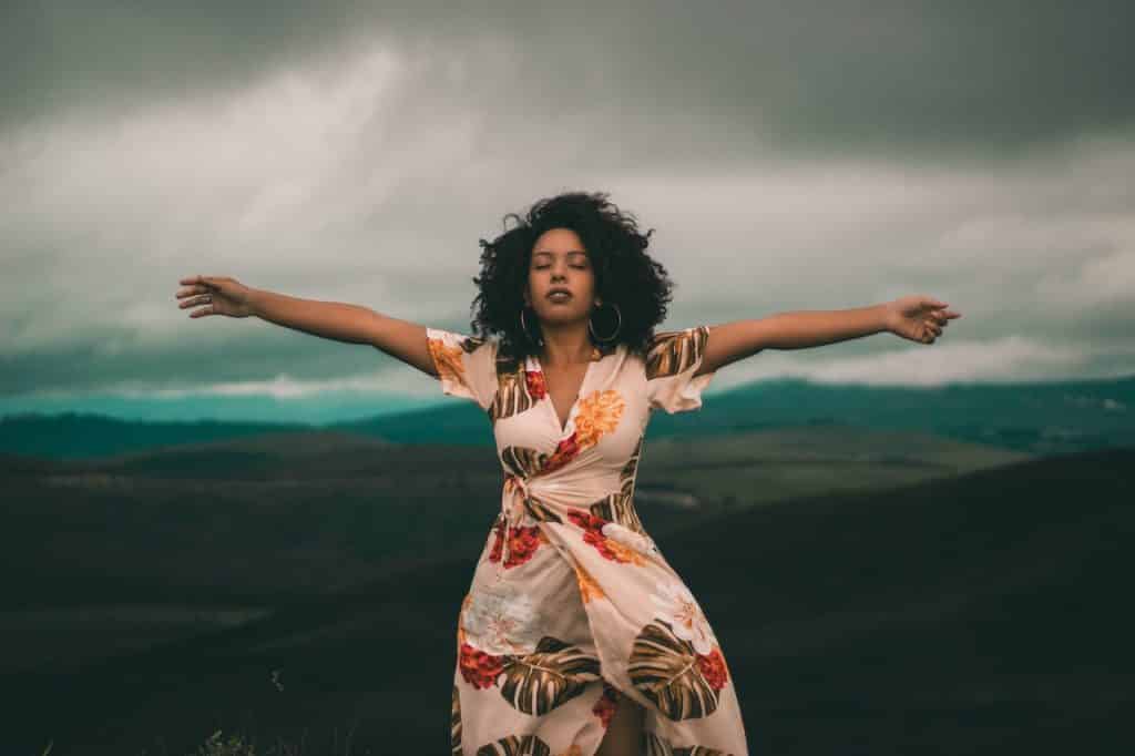 Mulher de cabelos cacheados com os braços abertos e os olhos fechados em um lugar alto, à frente de morros naturais.