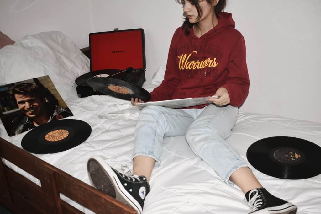 Mulher sentada na cama, com calça jeans e tênis All Star, em meio a discos de vinil.