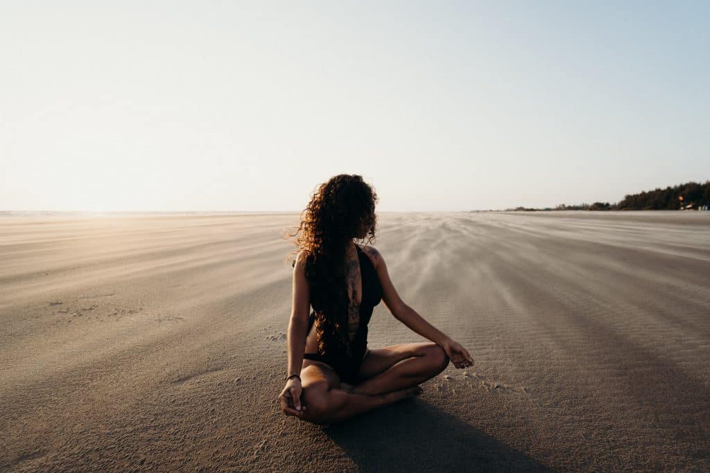 Mulher usando roupas de banho sentada na areia, meditando.