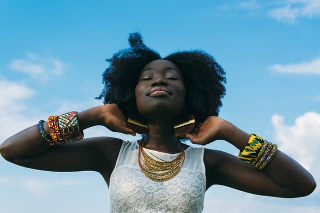 Mulher negra com mãos atras das orelhas e expressão de tranquilidade com céu azul ao fundo