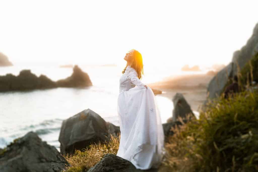 Imagem de uma mulher usando um vestido branco longo. Ela está de frente para o mar e está respirando o ar puro do local.
