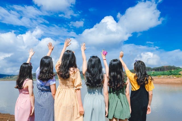 Mulheres de costas em frente lago com mãos para o alto e céu azul ao fundo