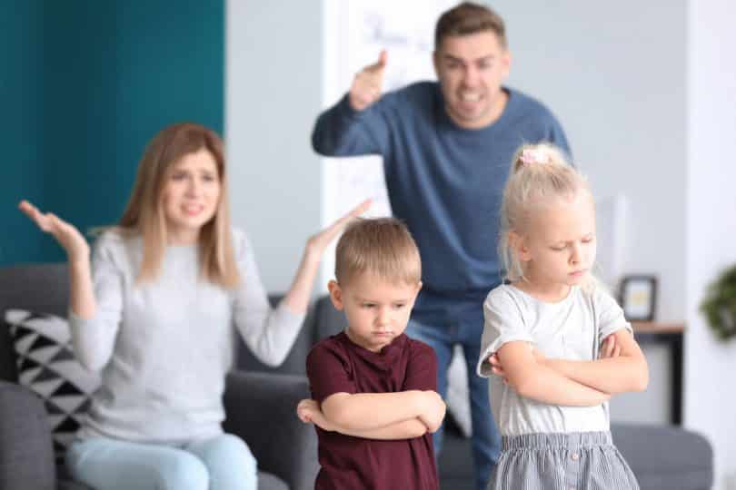 Pais gesticulando e gritando com seus dois filhos pequenos, que estão de braços cruzados.