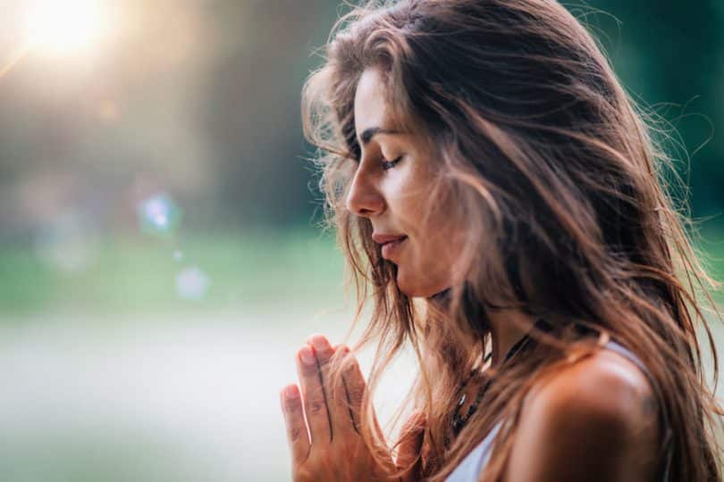 Mulher de cabelo solto e mãos em frente ao corpo com olhos fechados meditando