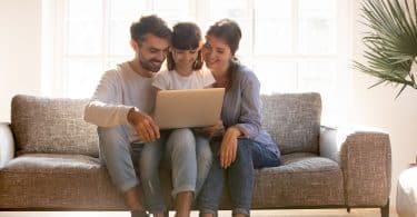 Mãe, pai e filha sentados no sofá, sorrindo e usando o computador.