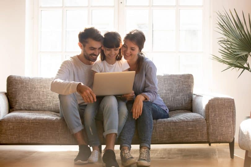 Mãe, pai e filha sentados no sofá, sorrindo e usando o computador.