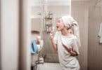 Mulher no banheiro cantando com uma toalha na cabeça e uma escova de dentes na mão.