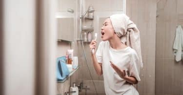 Mulher no banheiro cantando com uma toalha na cabeça e uma escova de dentes na mão.