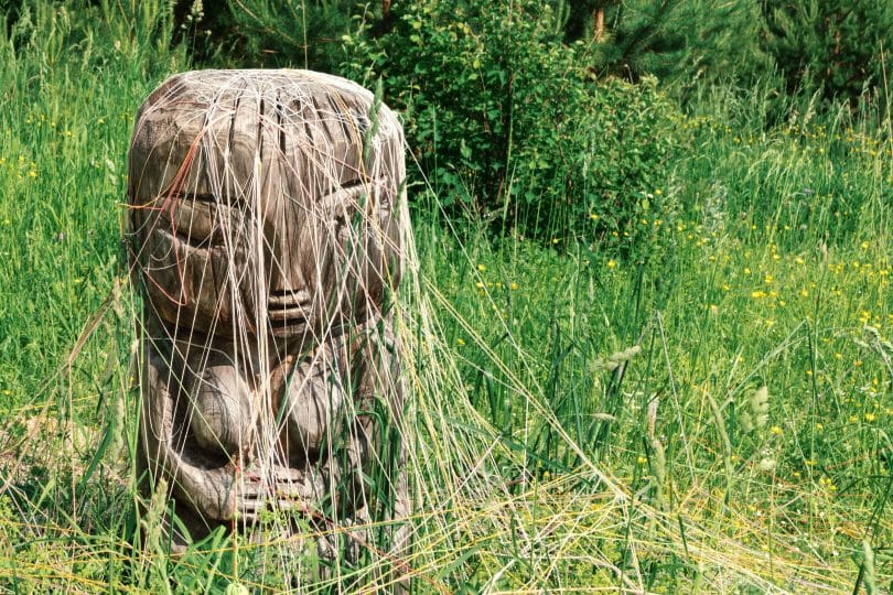 Imagem de um ser similar a uma mulher esculpida em pedra com cabelos que se misturam com as plantas do chão.