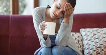 Mulher branca de olhos fechados sentada num sofá vermelho segurando uma caneca branca com uma mão e a outra levada à testa.