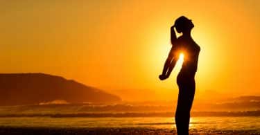 Mulher em pé se alongando na praia durante o pôr-do-sol