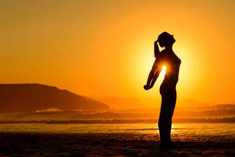 Mulher em pé se alongando na praia durante o pôr-do-sol