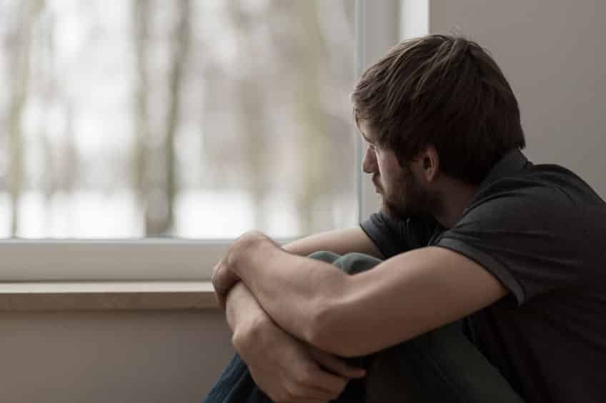 Homem olhando pela janela, sentado, com expressão triste.