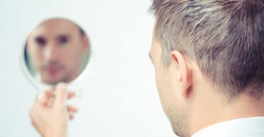 Homem de costas olhando o seu rosto refletido sem foco em um espelho redondo que está em sua mão.