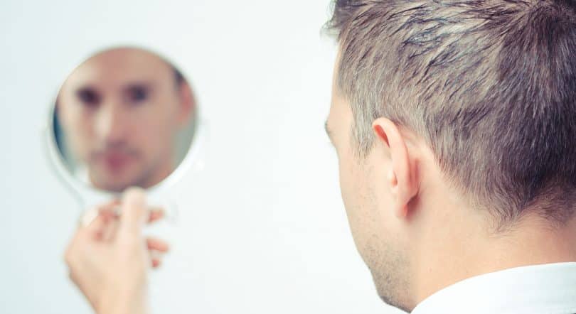 Homem de costas olhando o seu rosto refletido sem foco em um espelho redondo que está em sua mão.