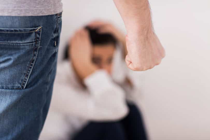 Imagem de uma mulher com medo, sentada no chão e com as mãos cobrindo o rosto. Em sua frente, aparece um homem com a mão fechada insinuando uma agressão física.