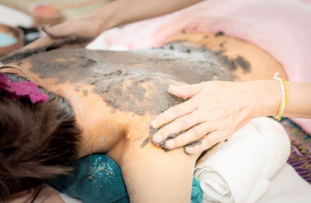 Mulher passando argila nas costas de outra mulher que esta em uma maca