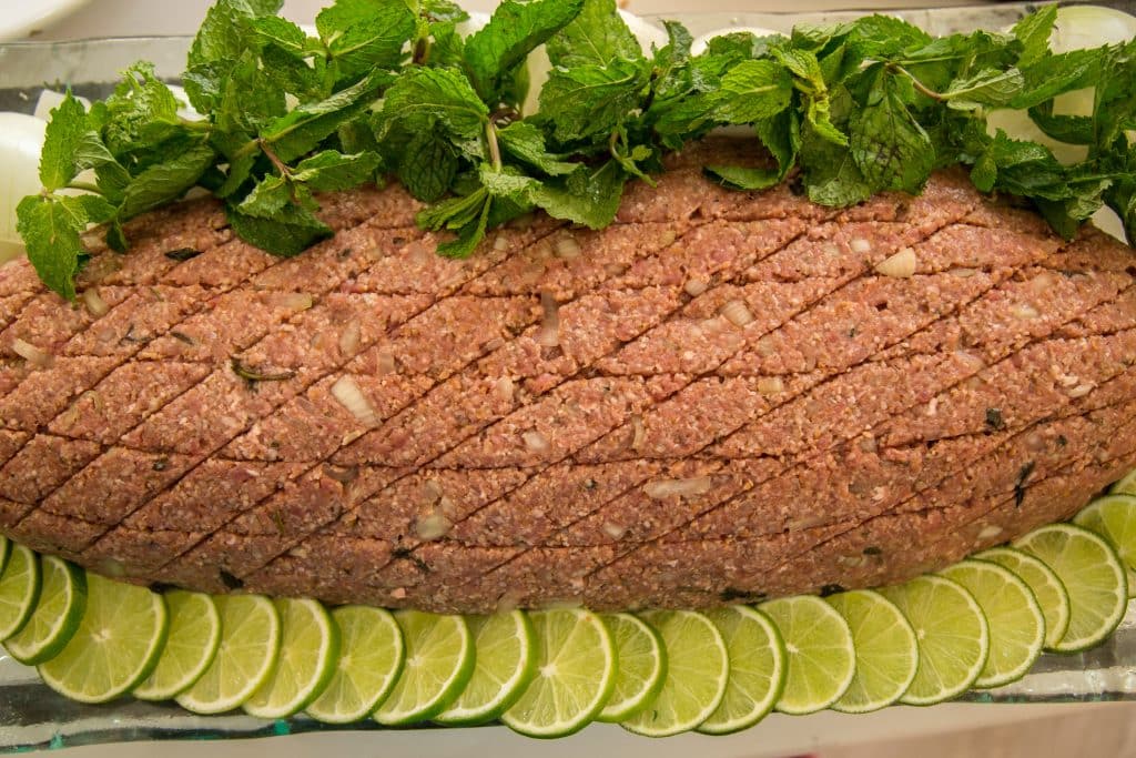 Imagem de uma travessa de quibe vegetariano pronto para ser assado. Ele está decorado com folhas de hortelã e rodelas de limão.