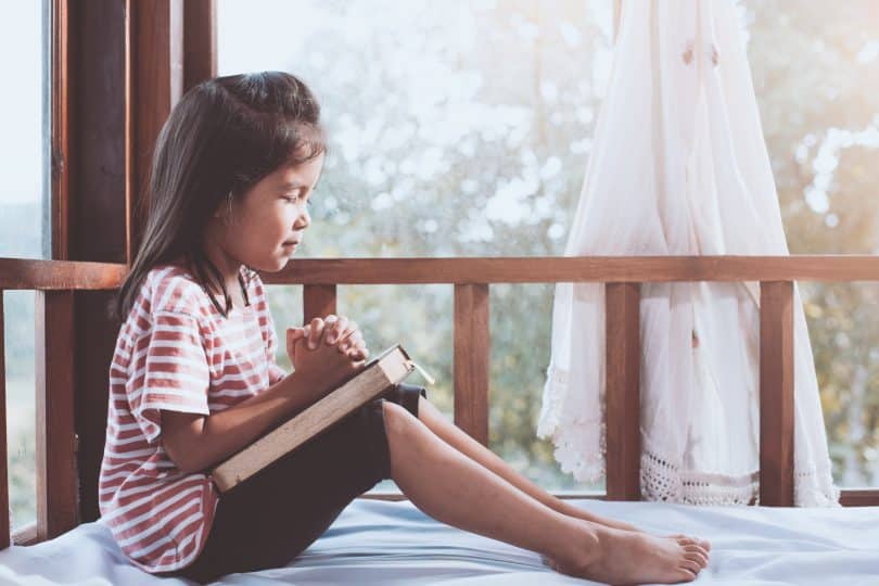 Menina pequena rezando em sua cama com janela ao fundo