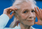 Mulher idosa de cabelos brancos com as mãos sobre o rosto, em fundo azul.