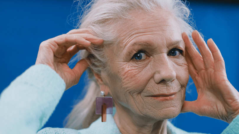 Mulher idosa de cabelos brancos com as mãos sobre o rosto, em fundo azul.