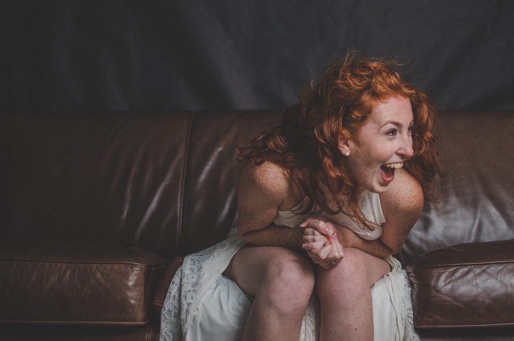 Imagem de uma linda mulher ruiva usando um vestido branco. Ela está sentada em um sofá de couro marrom e está rindo e com a autoestima elevada.
