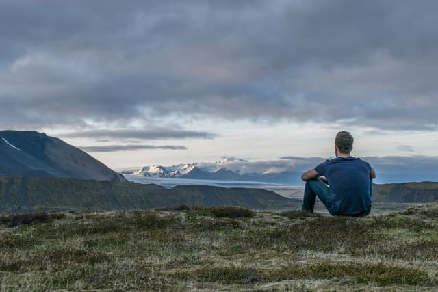 Homem sentado em montanha olhando para o céu e outras montanhas de costas