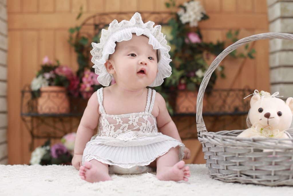 Imagem de uma linda bebê usando um vestidinho e uma touca de renda na cor branca. Ela está sentada sobre um tapete branco e ao lado dela uma cesta de vime com um ursinho dentro.