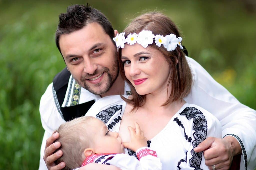 Imagem de um casal e o seu bebê. A mãe está amamentando o seu filho. Ela usa uma tiara de flores e todos estão felizes.
