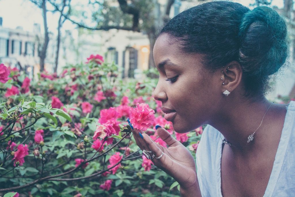 Imagem de uma linda mulher negra, feliz e com a autoestima elevada. Ela está cheirando uma linda flor cor de rosa.
