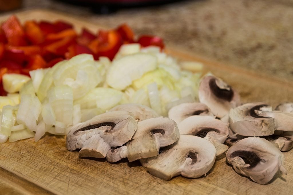 Cogumelos, cebolas e pimentões picados em uma tábua.