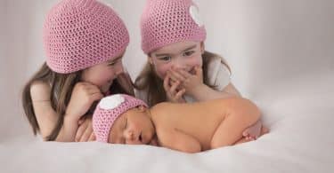 Imagem de três meninas, sendo que uma delas é uma recém nascida. As três usam uma touca rosa de lã.