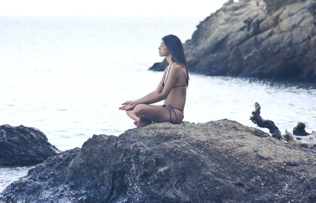 Mulher sentada em uma pedra da praia. Ela está em posição de meditação.