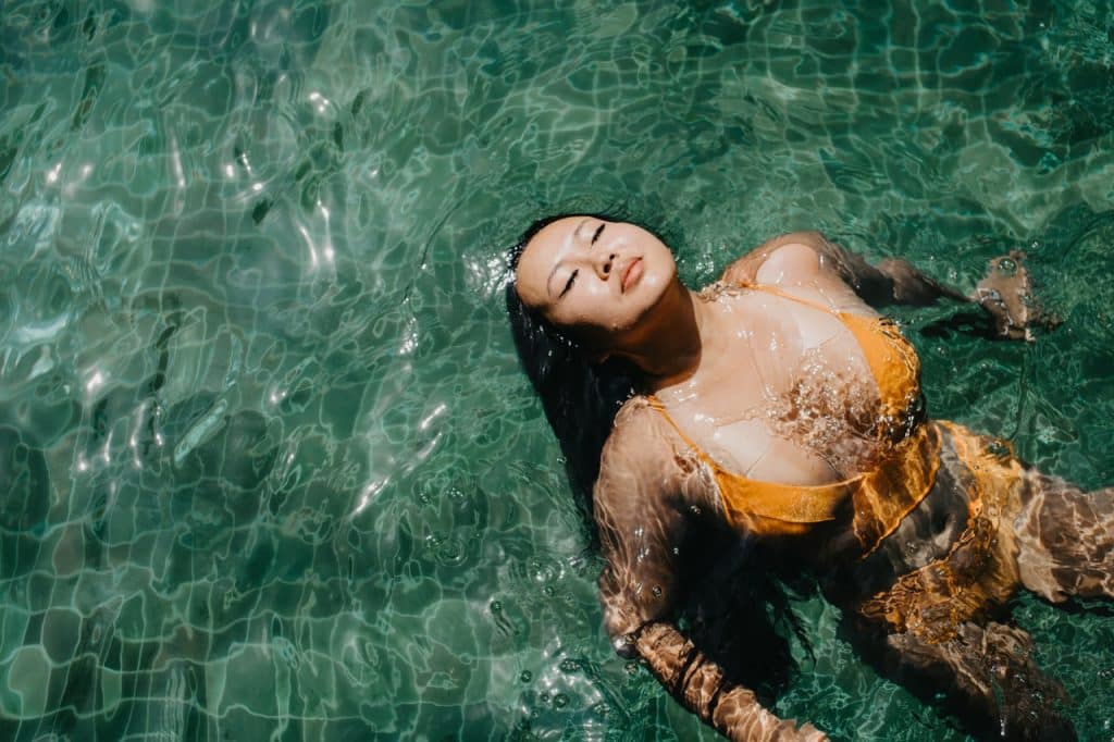 Mulher de cabelos longos na piscina, com a cabeça para cima e os olhos fechados.