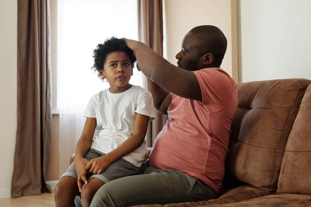 Pai arrumando o cabelo do filho enquanto ambos conversam no sofá.