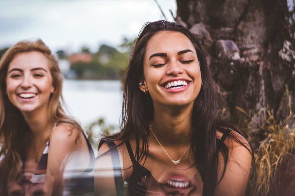 Duas mulheres sorrindo de olhos fechados ao lado de uma árvore.