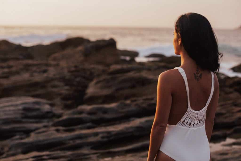 Mulher branca com maiô branco de costas observando a praia