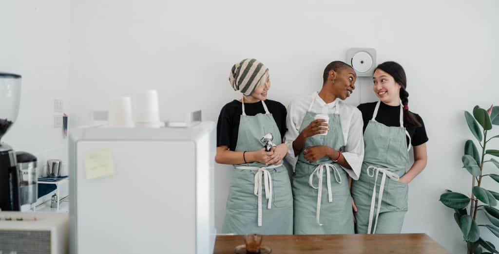 Três mulheres conversando lado a lado, usando aventais de culinária.