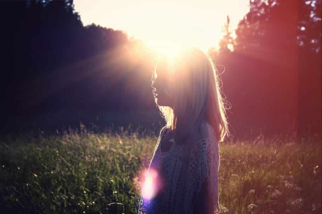 Imagem de uma mulher de lado em meio à um campo verde com grande reflexo do sol.