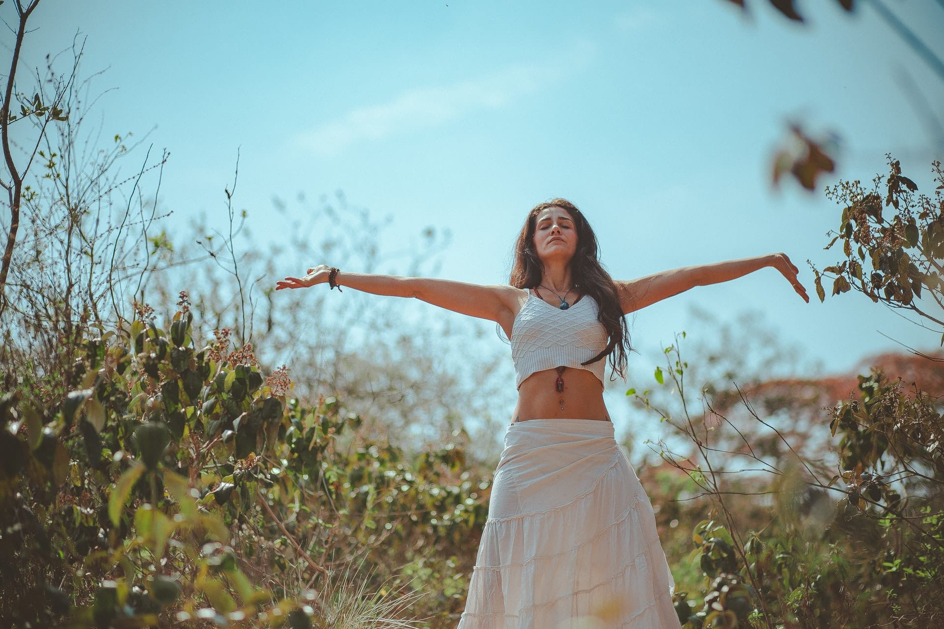 Mulher branca meditando com braços abertos ao ar livre.