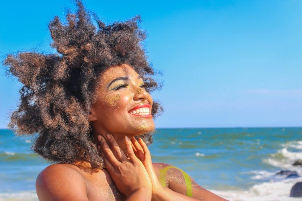 Mulher vista de perfil, na praia, sorrindo com seus cabelos cacheados ao vento