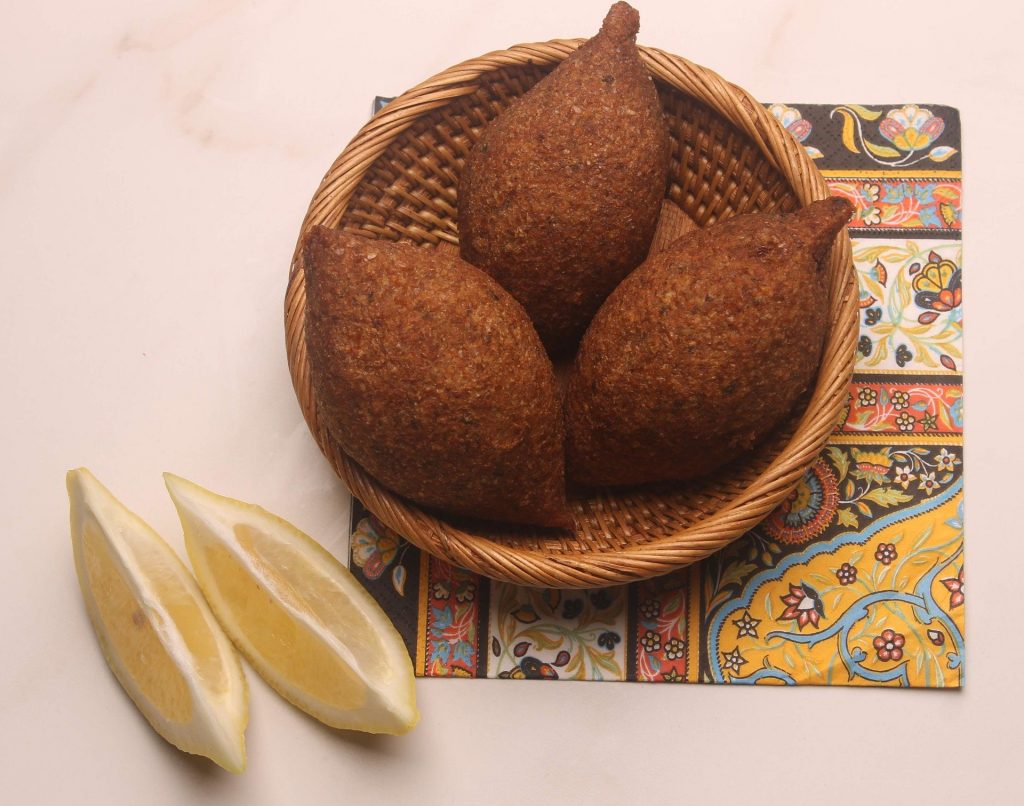 Imagem de três quibes vegetarianos prontos para serem servidos. Eles estão sobre uma cestinha de vime e ao lado um limão cortado em fatias.