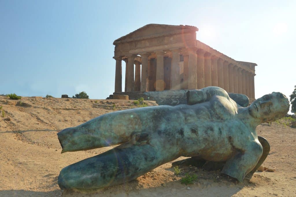 Estátua de Ícaro caída no chão, sem as pernas, braços e com as asas quebradas, em frente ao Partenon - um monumento cheio de pilares.