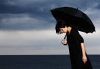 Imagem de um dia nublado na praia. Ao fundo uma mulher usando uma roupa preta e segurando um guarda chuva preto. Ela está com depressão.