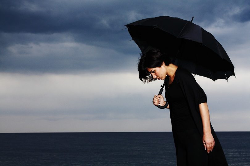 Imagem de um dia nublado na praia. Ao fundo uma mulher usando uma roupa preta e segurando um guarda chuva preto. Ela está com depressão.