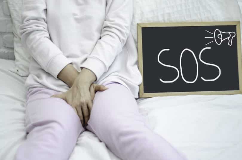 Imagem de uma mulher sentada sobre a cama. Ela usa um moletom, está com sintomas de candidíase. Ao lado dela uma lousa escrita SOS.