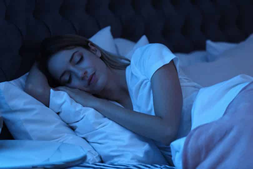 Mulher jovem dormindo em uma cama, em um ambiente com luz baixa de tom azul.