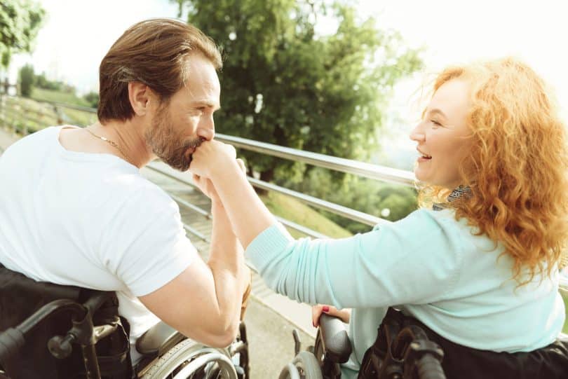 Homem e mulher brancos, ambos em cadeira de rodas, o homem beijando a mão da mulher.