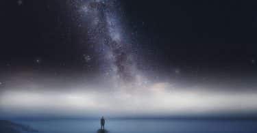 Silhueta de um homem em cima de uma pedra no meio do mar com um céu estrelado acima dele.